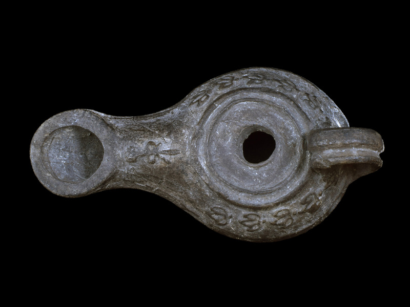 Sharply carinated reservoir w flat discus w fairly small fill hole  surrounded by two raised ridges, bipartite high loop handle fr carination to edge of discus, nozzle w large circular wick hole.  Shoulder decorated w relief  band of ivy leaves & surface of nozzle w ?ax tied w ribbon.  Now all dark gray, ?paint and/or firing.