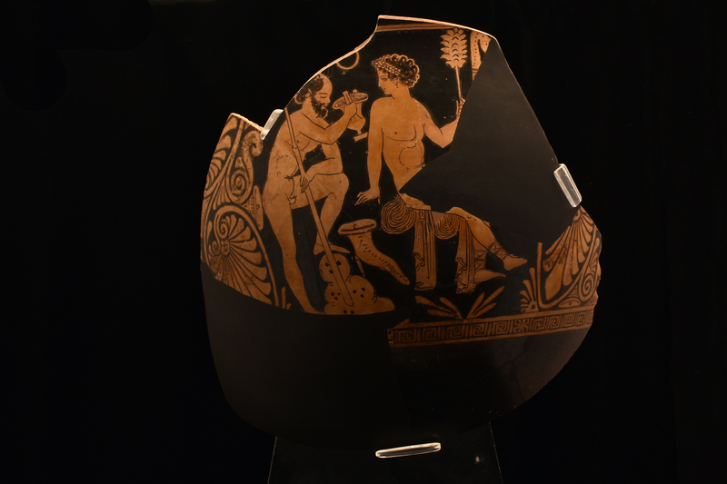 Large fragment from Side A w Dionysus sitting on rock over which his cloak draped, .body in 3/4 view to his l, but w head turned in profile to mature satyr drinking from kantharos who stands behind him, his l leg raised on rock; elaborate multiple palmette & volute elements flank scene.  Effeminate nude Dionysus w broad wreath over short curly locks defined by reserve border, dangling earring & Thracian boots; r arm leaning back on rock, l hand raised holding thyrsus.  Satyr, nude w r arm raised to hold kantharos, l arm resting on bent l leg; curly hair, but head balding, pointed ears.  Limited anatomical details.  Drinking horn on ground between figures & dotted & fringed scarf hanging in field next to thyrsus ; floral leaf device below Dionysus.  Above scene narrow reserved band, below maeander band broken intermittently w star device.