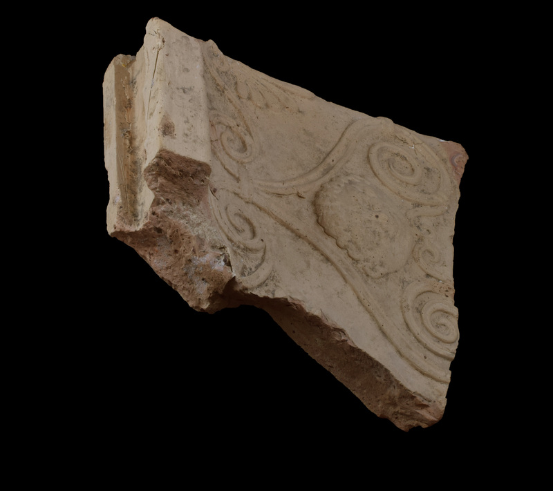 Flat slab with overhanging cavetto molding, top surface flat with broad deep rectangular-sectioned groove.  On exterior surface relief decoration: double scroll composed of scrolls at top small and bound together with horizontal band, scrolls below widely separated with Gorgoneion in space between; beneath head small double scroll the termination of bands from large scrolls above.   At top curving bands extend from base of scrolls to either side and terminate in inverted palmettes