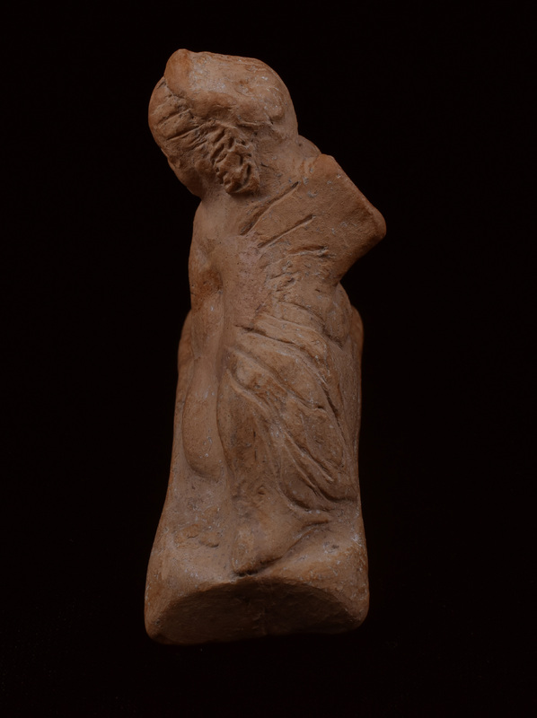 On rectangular base chubby Eros reclining on right elbow with right hand on rabbit.  Round face with eyebrows, eyes, nostrils, mouth, and cleft in chin articulated by incision; wreath on head with hair articulated by incisions going from forehead down back of head on either side of wreath, with clumps of hair articulated by irregular incision falling on either side of head.  Left arm covered by drapery stretched out and resting on bent left leg; incised lines roughly articulating folds of drapery; some articulation of feathers of left wing.  Raised band runs around back of figure under back of wings, possibly meant as drapery.