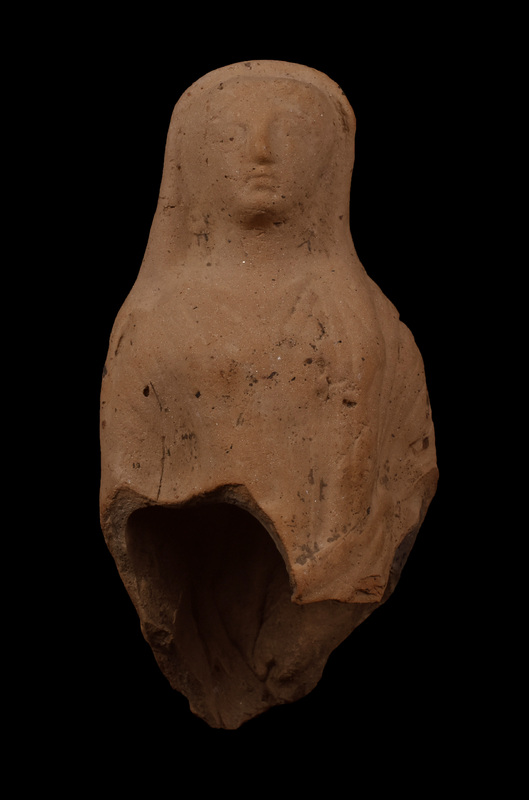Mouldmade standing figure w himation draped over head and falling down over outside of r arm, coming forward over l breast, w fold coming across fr r side and tucked under l arm folds of chiton and himation articulated by incision.  Hairline, eyes, nose, and mouth articulated by incision.