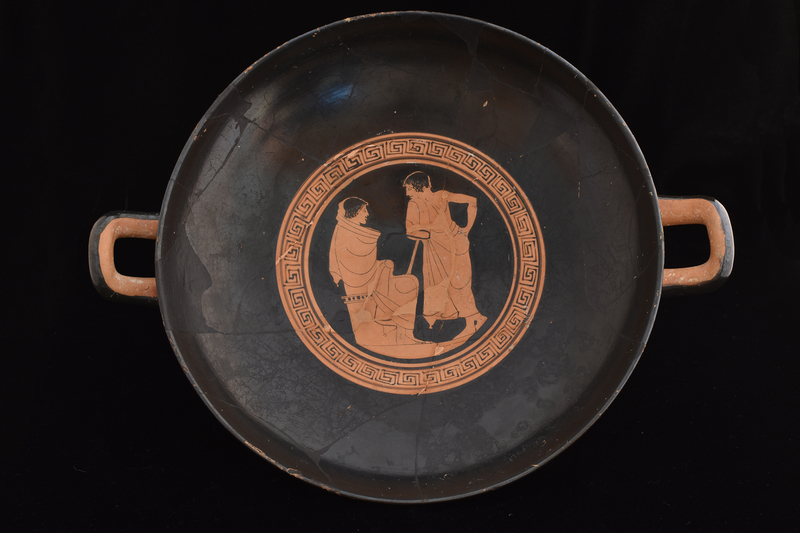 Kylix by Sabouroff Ptr, v. Oakley letter in file.  Decorated w scenes fr palaestra w  palmettes under handles; interior of handles & adjacent body, edge of foot & int of stem reserved. Tondo defined by discontinuous maeander w reserved exerque: Youth at l seated on altar facing r, swathed in himation which pulled over head on which added red fillet; at r standing youth in 3/4 view fr rear, r hand on hip, leans toward first, resting on smooth staff under l arm; wreath in added red.  Side A: Presentation of fillet to lyre-player; fr l to r: youth w fillet in added red in himation leaning r w staff under l arm & holding fillet in added red toward lyre-player swathed in himation, who sits on altar facing r holding lyre upright and wreath in added red; to r of lyre player frontal youth in himation pulled over l shoulder leaving chest bare,  r arm bent w hand on hip and w staff under l arm, face in profile to l, wreath in added red, feet crossed at ankles; at l another youth in himation, 3/4 view fr back, leaning in on staff under l arm  toward center, w r arm bent & hand on hip, face in profile to l w wreath roughly incised, possible modern;  on background aryballoi, strigils and one wooden cross; K A L O S in added red between figures 1 and 4.  Side B: Four youths in conversation fr l to r: frontal youth holding staff w curved top in r hand, r arm bent under himation, face in profile to r, wreath in added red; youth swathed in himation seated on altar facing r w arms bent holding bent r knee, all under himation; youth in 3/4 view fr rear leaning l w arm bent & hand on hip, leaning on staff under l arm, face in profile to l, fillet in added red; frontal youth with arms swathed in himation, r raised, left bent to hip, face in profile to l; on backgrd aryballoi, flute cases, strigils.