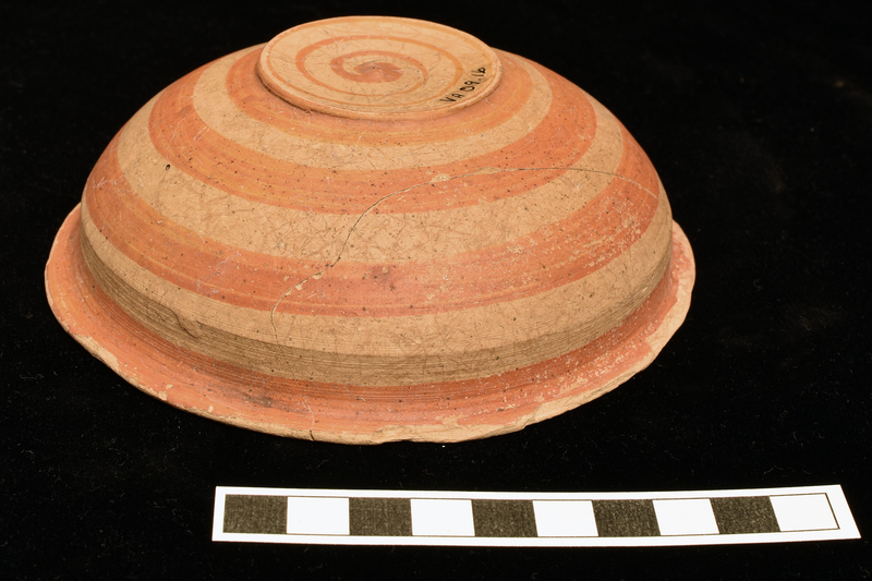 Shallow bowl with convex sides rising to poorly articulated carination and continuing  vertically to sharply outturned rim; low off-set base shallowly concave on underside.  Decorated interior and exterior with broad stripes in orange paint: int, solidly painted circle on base, single stripe about mid-body and narrow stripe on interior of rim which continues on to top and underside of rim: ext, 4 stripes from underside of rim to and including base, concentric swirl of paint on underside of base ending in irregular circle.  Two holes close to one another at juncture of rim and body.