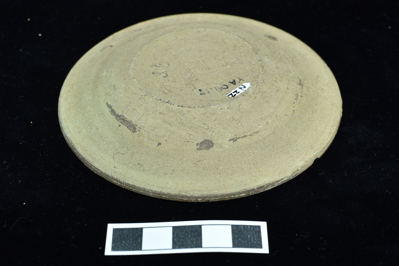 Small impasto dish with broad shallowly convex rim. Upper surface solidly coated on rim with paint continuing as broad band on upper wall of dish proper  which continues as spiral which ends in center of interior.  On underside traces of stripe just below rim and two dots just above stripe at one quarter of circumference.