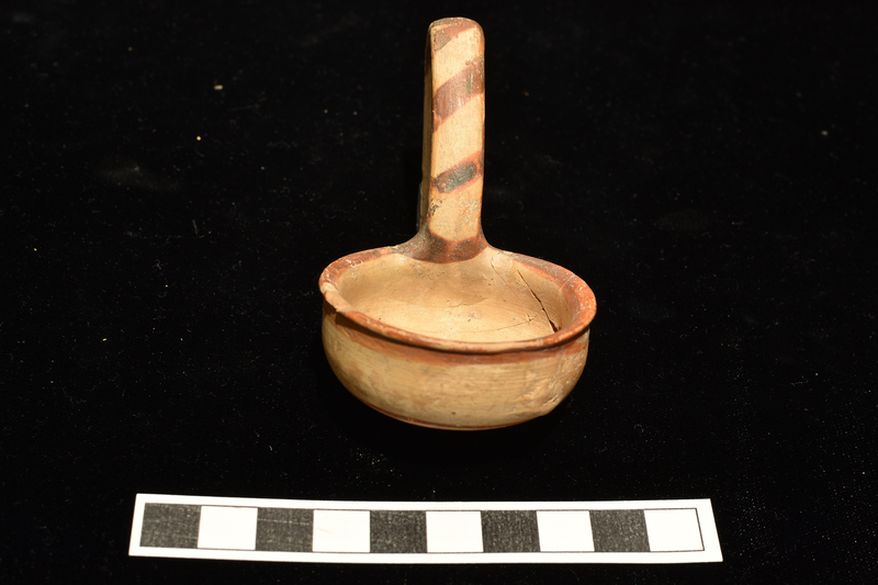 Bowl with shallowly convex sides which rise to small outturned rim, rounded articulation with convex base, nippled in center.  High swinging handle of flattened elliptical section from juncture of body and base to top of rim.  Decorated on exterior and handle: band on rim continuing as stripe on upper body, four narrow concentric stripes at outer edge of base, central nipple solidly coated; handle on surface which faces away from bowl solidly coated except for deep V-shaped area at base, diagonal stripes on surface which faces towards bowl.