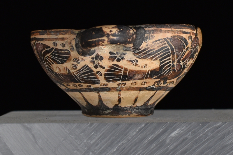 Bowl w convex sides spreading from low offset base to plain rim, prominent nipple on int center; horizontal handles of rounded rectangular section.attached at either side at rim.  Main zone of decoration defined by 2 h stripes above, one below filled w opposing sphinxes on opposite sides of inverted palmette w small blob rosettes in main fields, larger under handles, all decorated w fine incised detail; below rays rise from base which solidly coated on ext w paint continuing onto to underside of base; handles solidly coated on ext.; int thinly coated w paint of varying thickness, so as to produce h stripes.