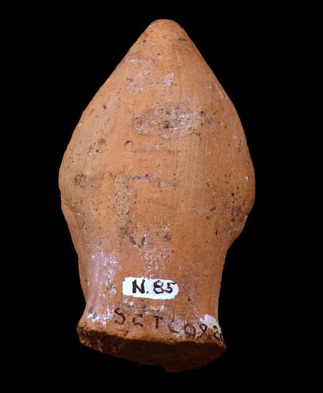 Head of male in pointed cap.  Face triangular with round blunted chin, pellet eyes under overhanging brows, nose modeled, mouth indicated by horizontal incision, ears indicated by small scallop-shape attachments.