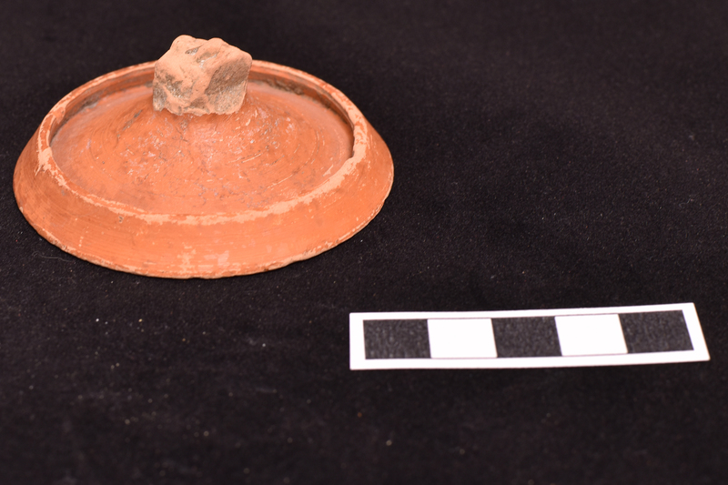 Lid, probably for small jar, concave below with raised edge encircling convex upper surface from which handle rises; prominent wheel marks on upper surface and on under surface single incised line close to edge and another encircling concavity.  Slipped all over.