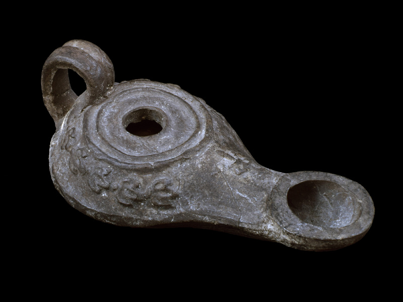 Sharply carinated reservoir w flat discus w fairly small fill hole  surrounded by two raised ridges, bipartite high loop handle fr carination to edge of discus, nozzle w large circular wick hole.  Shoulder decorated w relief  band of ivy leaves & surface of nozzle w ?ax tied w ribbon.  Now all dark gray, ?paint and/or firing.