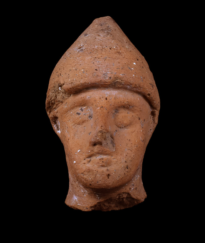 Head of male in pointed cap.  Face triangular with round blunted chin, pellet eyes under overhanging brows, nose modeled, mouth indicated by horizontal incision, ears indicated by small scallop-shape attachments.