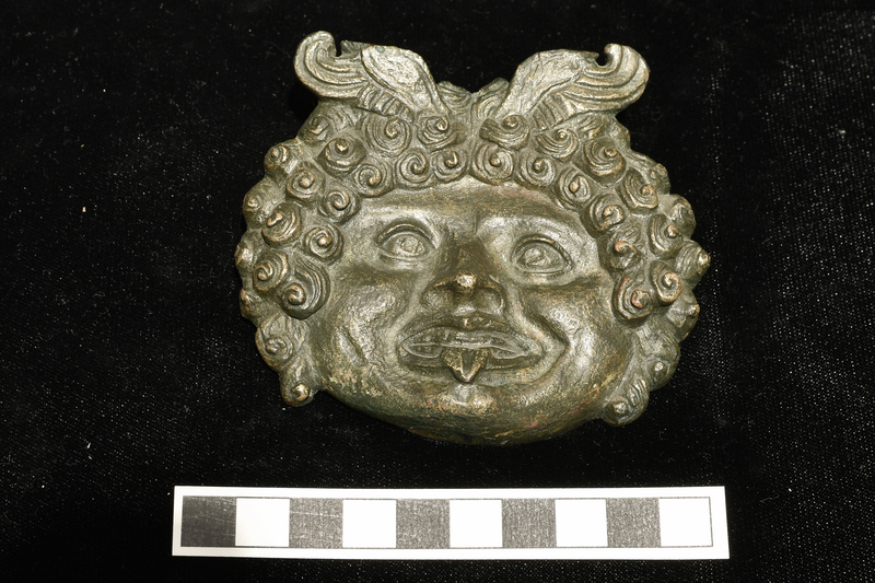 Oval plaque with face surrounded by mass of snail-shell curls with hair strands articulated; at part in center bipartite wings with feathers articulated in outer portion rise diagonally to the sides.  Eyes and mouth clearly articulated with raised contours for lids and lips; circular depressions in pupils of eyes; between lips incised line marking juncture of teeth, which are separately articulated, with triangular tongue projecting over center of lower lip.  Forehead slightly depressed, cheeks round and projecting, and area around mouth recessed with projecting chin with central cleft.
