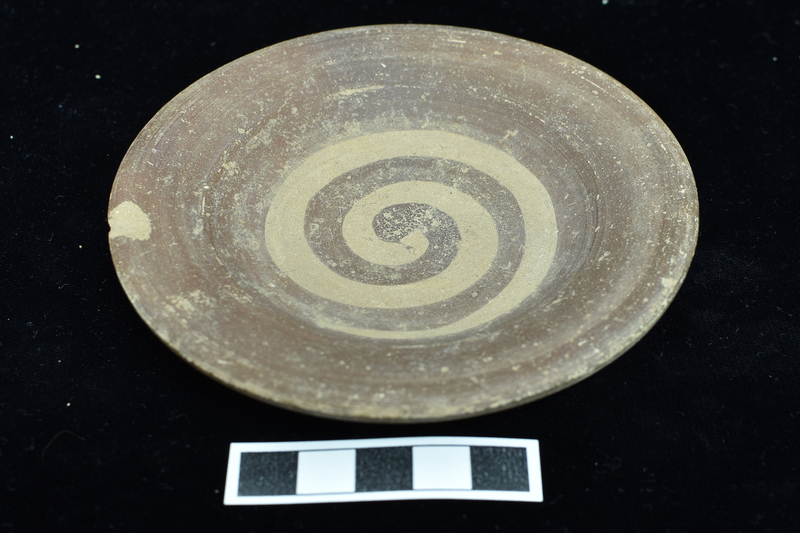 Small impasto dish with broad shallowly convex rim. Upper surface solidly coated on rim with paint continuing as broad band on upper wall of dish proper  which continues as spiral which ends in center of interior.  On underside traces of stripe just below rim and two dots just above stripe at one quarter of circumference.