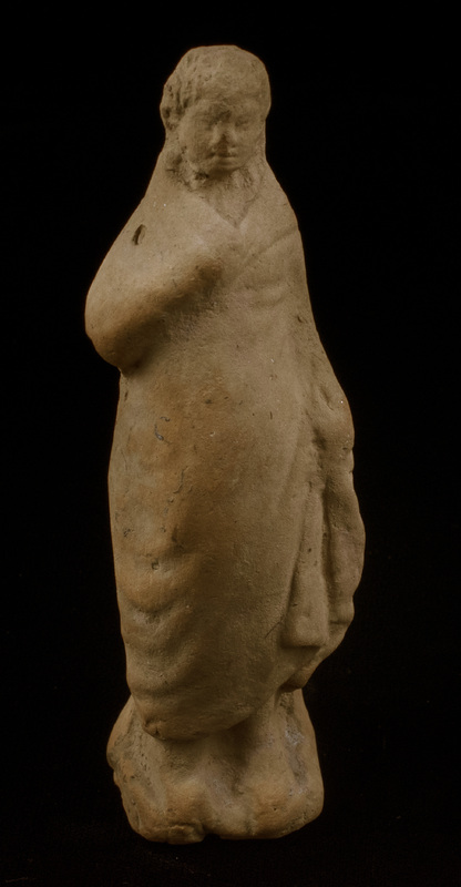 Mouldmade standing figure of Sophoclean type with right arm and hand completely covered by himation and left arm, slightly bent, close to side with large clump of drapery falling forward from left hand; left foot projecting slightly before right, small base.  Eyes, mouth and nose articulated and irregular incisions articulating hair mass which is particularly defined around face.  Very large vent hole in plain back.