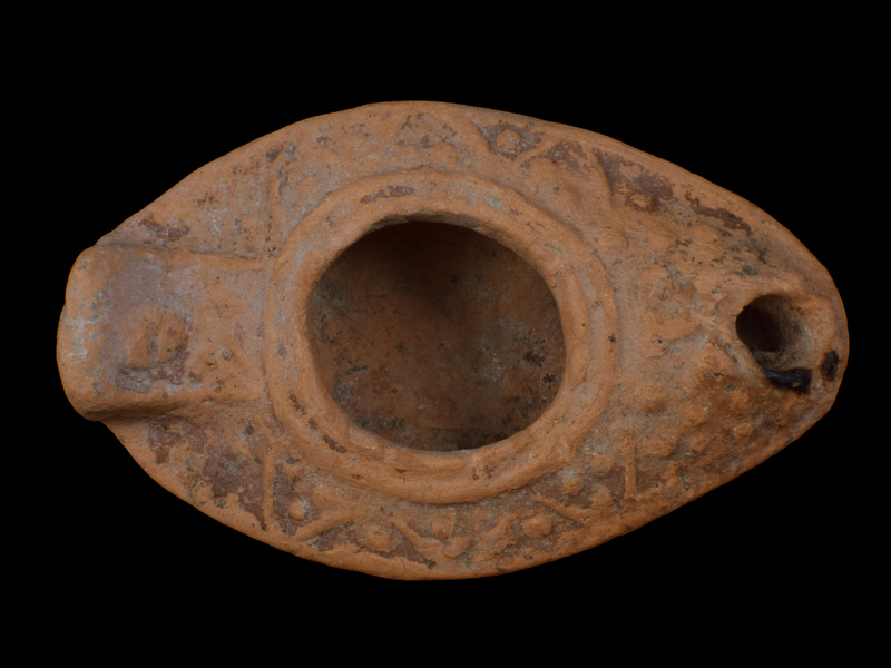 Slipper lamp w upturned solid rectangular lug handle, edges of which defined by relief band within which relief X below, relief ?rosette above.  Carinated reservoir w flattened carination, large fill hole surrounded by two relief bands;  on upper surface decoration of open V's pointing toward carination within and between which relief dots; relief Greek cross on nozzle.  Ca flat base depressed within poorly articulated raised band; possible mark at edge of band two indistinct depressions.  Top surface coated w red paint, some of which dripped to underside; blackened nozzle.