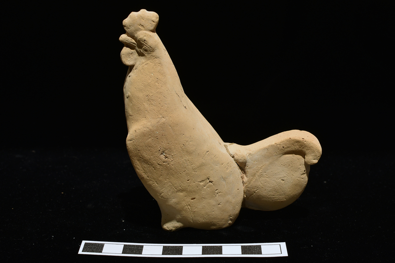 Standing rooster w very short front legs and vestigial haunches providing squarish base.  On head crest, beak, wattles & arms articulated; wings indicated by ridge on either side of body; tail a vertical semicircular projection attached to top of which a curving plume.