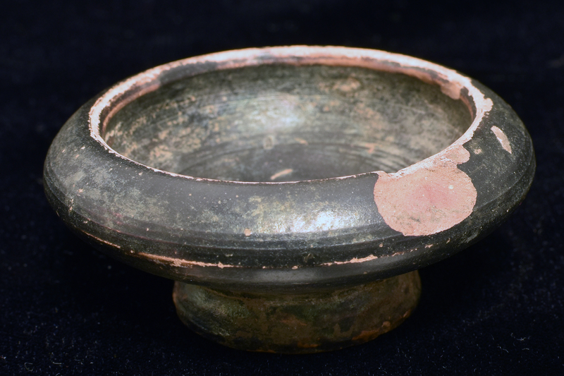 Squat carinated bowl on low pedestal foot with nippled center on underside; plain rim.  Impressed decoration on int center:line of 3 raised dots in oval w single dots on either side of center dot in cruciform configuration.  Coated all over w dark paint.