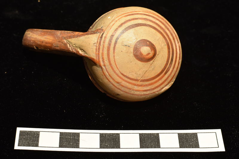 Bowl with shallowly convex sides which rise to small outturned rim, rounded articulation with convex base, nippled in center.  High swinging handle of flattened elliptical section from juncture of body and base to top of rim.  Decorated on exterior and handle: band on rim continuing as stripe on upper body, four narrow concentric stripes at outer edge of base, central nipple solidly coated; handle on surface which faces away from bowl solidly coated except for deep V-shaped area at base, diagonal stripes on surface which faces towards bowl.