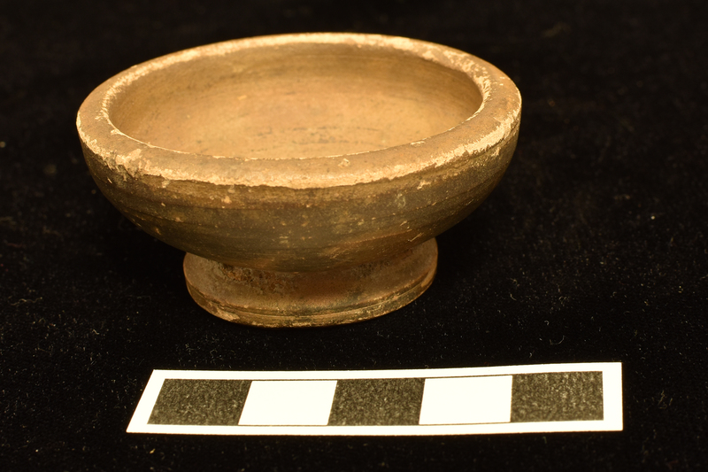 Bowl w convex sides spreading fr low pedestal base, underside flat w tiny applied pellet in center, to thick, plain horizontal rim.  Coated all over int & most of ext w dark paint, except for small portion of base, large splash of paint on underside of base.