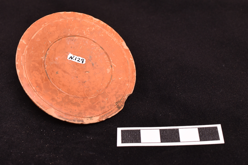 Lid, probably for small jar, concave below with raised edge encircling convex upper surface from which handle rises; prominent wheel marks on upper surface and on under surface single incised line close to edge and another encircling concavity.  Slipped all over.