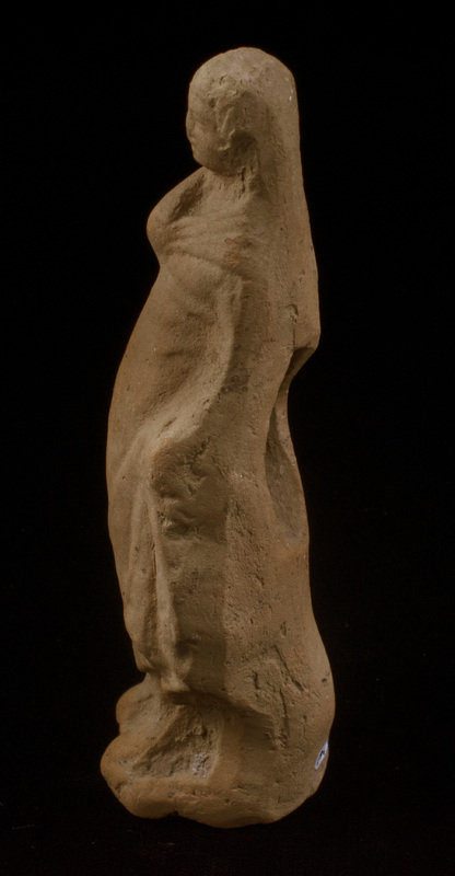 Mouldmade standing figure of Sophoclean type with right arm and hand completely covered by himation and left arm, slightly bent, close to side with large clump of drapery falling forward from left hand; left foot projecting slightly before right, small base.  Eyes, mouth and nose articulated and irregular incisions articulating hair mass which is particularly defined around face.  Very large vent hole in plain back.