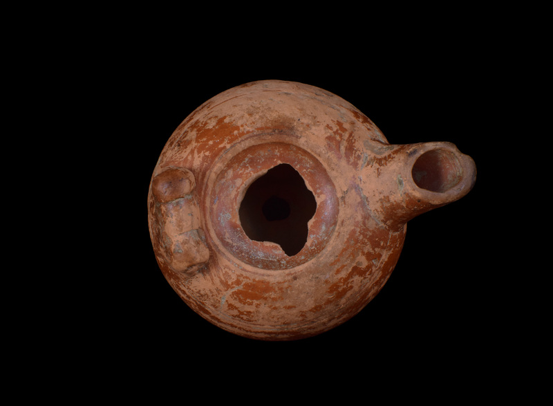 Squat round-carinated body on sharply defined offset base; shallowly concave discus w finely incised line defining it on ext, pairs of similar incised lines on body at mid-nozzle & at base of nozzle.  Opposite nozzle on shoulder horizontal volute handle.  Coated on ext w glossy red slip which darkened at edge of nozzle.  Scratched in base uppercase Alpha,  Gamma, Iota.