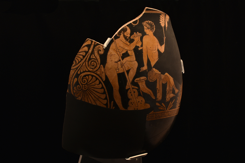 Large fragment from Side A w Dionysus sitting on rock over which his cloak draped, .body in 3/4 view to his l, but w head turned in profile to mature satyr drinking from kantharos who stands behind him, his l leg raised on rock; elaborate multiple palmette & volute elements flank scene.  Effeminate nude Dionysus w broad wreath over short curly locks defined by reserve border, dangling earring & Thracian boots; r arm leaning back on rock, l hand raised holding thyrsus.  Satyr, nude w r arm raised to hold kantharos, l arm resting on bent l leg; curly hair, but head balding, pointed ears.  Limited anatomical details.  Drinking horn on ground between figures & dotted & fringed scarf hanging in field next to thyrsus ; floral leaf device below Dionysus.  Above scene narrow reserved band, below maeander band broken intermittently w star device.